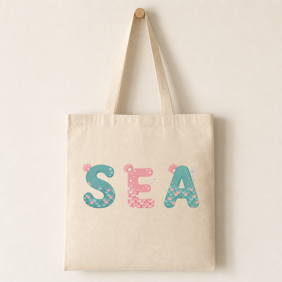 Canvas tote bag mockup printed with a mermaid alphabet letter featuring scallop scales, a pale pink shell at the top-left and small bubbles.